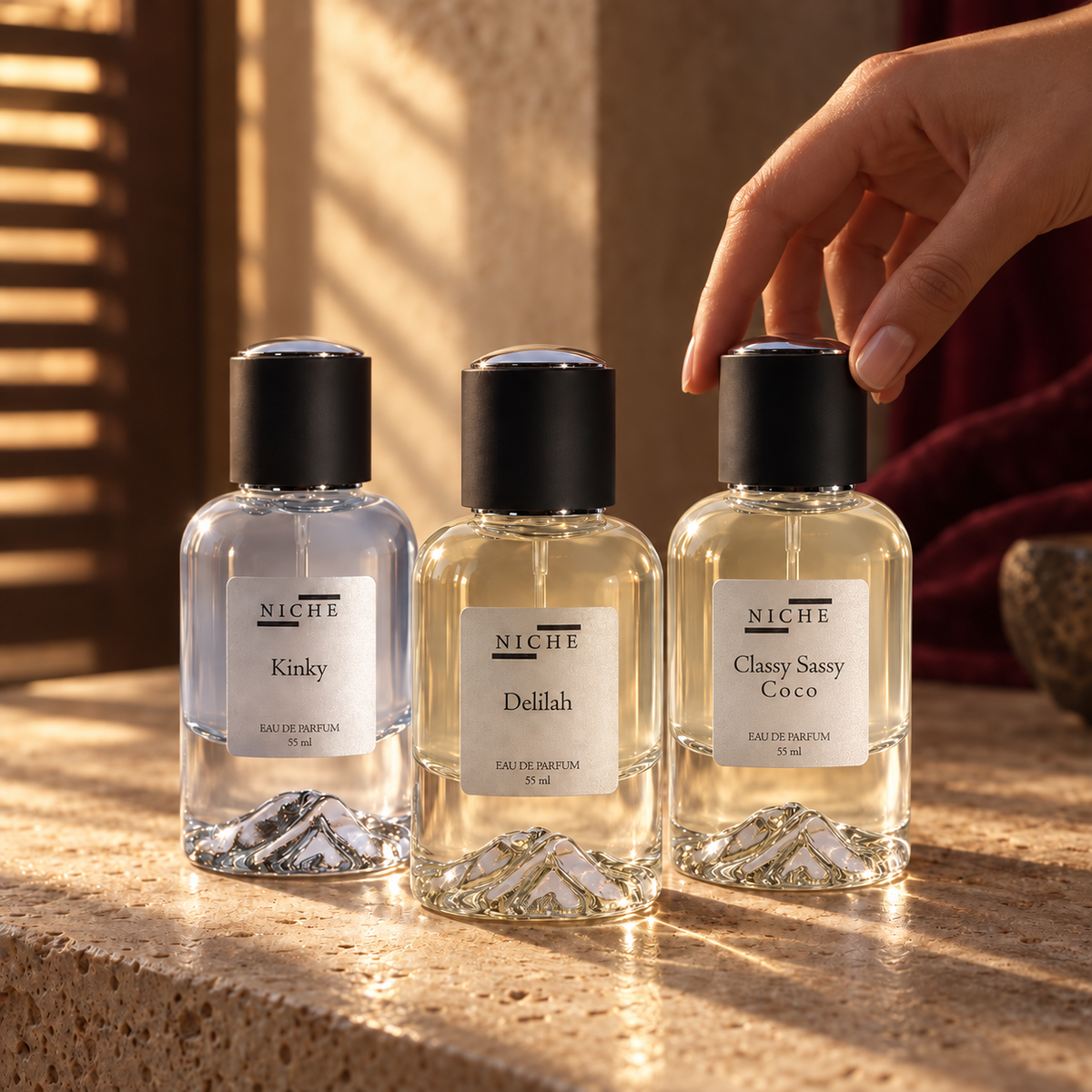 Kinky, Delilah and Classy Sassy Coco fragrances in warm light, with a hand selecting a bottle